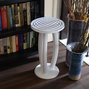 May include: A white pedestal table with a circular top featuring a black grid pattern. The table has three curved legs that connect to a round base. The design is simple and functional, providing a stable surface for display or utility.