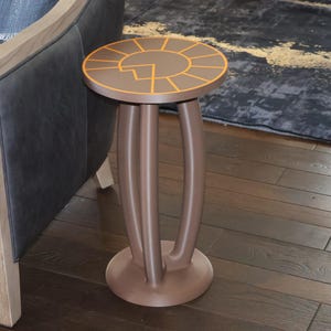 May include: A brown circular side table featuring a three-legged base and a flat top surface. The tabletop is decorated with a geometric sun-like pattern outlined in thin orange lines, creating a segmented design across the circular surface.