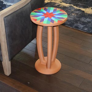 May include: A round side table with a matte terracotta-colored base and three curved legs. The tabletop features a vibrant, multi-colored geometric star pattern in shades of red, blue, green, and purple, creating a bold, symmetrical design.