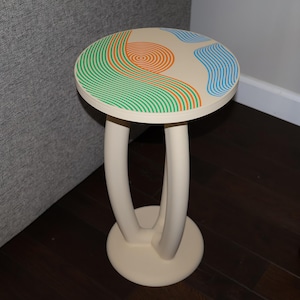 May include: A round side table with a cream-colored base and two curved legs. The tabletop features a cream background with a graphic design of concentric orange circles, wavy green lines, and blue shapes arranged in a landscape-like pattern.