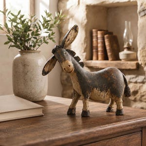 May include: A standing donkey figurine with a distressed finish in shades of dark grey, brown, and cream. The sculpture features long, pointed ears, a short mane, and a small tail, showcasing a weathered, textured appearance throughout its body.
