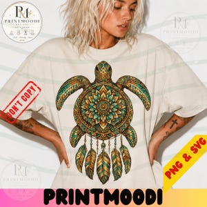 May include: A beige short-sleeved t-shirt featuring a large graphic of a sea turtle with a mandala-patterned shell in shades of teal, gold, and brown. The turtle design includes dangling feathers hanging from the bottom of the shell.