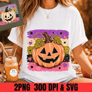 May include: A white t-shirt featuring a central graphic of an orange jack-o'-lantern pumpkin with green leaves, set against a background of horizontal purple, beige, green, and pink brushstrokes decorated with black stars and small pink hearts.
