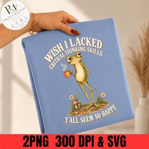 May include: A light blue t-shirt featuring a graphic of a green frog holding an orange mug, surrounded by small mushrooms and flowers. The text reads, "WISH I LACKED CRITICAL THINKING SKILLS Y'ALL SEEM SO HAPPY" in a curved, bold, cream-coloured font.