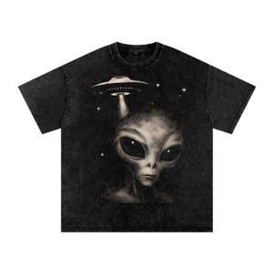 Op de afbeelding: Een zwart acid-wash t-shirt met een grafische print van een alienhoofd met grote donkere ogen en een vliegende schotel erboven. Het ontwerp is uitgevoerd in een halftoon-stippenpatroon met kleine vierpuntige sterren verspreid rondom het onderwerp.