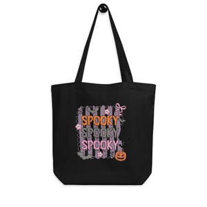 Könnte beinhalten: Eine schwarze Tragetasche mit einem Grafikdruck, der den Text SPOOKY dreimal in Orange, Grau und Pink wiederholt. Das Design enthält Illustrationen von Fledermäusen, einem Kürbis, kleinen Blumen, einer rosa Schleife und einem Spinnennetz auf Streifen.