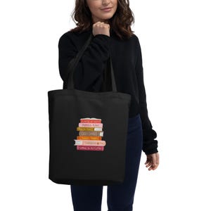Könnte beinhalten: Eine schwarze Tragetasche mit einem Grafikdruck eines vertikalen Stapels bunter Bücher. Die Buchrücken zeigen Text, darunter Pumpkin Plans, Cozy Chaos, Thanksgiving Pies und Letters to Autumn, zusammen mit verschiedenen herbstlichen Farben und Symbolen.