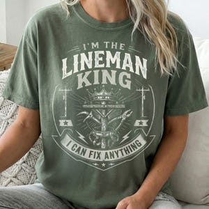 May include: An olive green short-sleeve t-shirt featuring a white graphic print. The design includes the text I'M THE LINEMAN KING and I CAN FIX ANYTHING, surrounding an illustration of a utility pole, a crown, and various lineman tools.