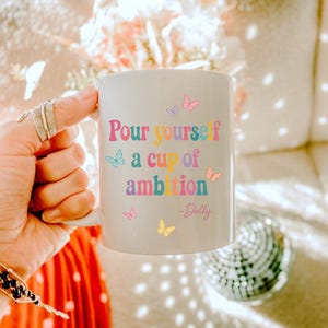 May include: A white ceramic mug featuring the colorful text "Pour yourself a cup of ambition" followed by "-Dolly" in a script font. The design is decorated with small, multi-colored butterflies in shades of pink, blue, yellow, and purple.