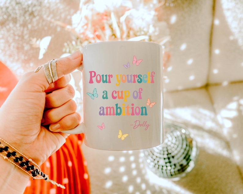 May include: A white ceramic mug featuring the colorful text "Pour yourself a cup of ambition" followed by "-Dolly" in a script font. The design is decorated with small, multi-colored butterflies in shades of pink, blue, yellow, and purple.