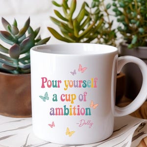 May include: A white ceramic mug featuring the colorful text "Pour yourself a cup of ambition" followed by "-Dolly" in a script font. The design is decorated with small, multi-colored butterflies in shades of pink, yellow, blue, and purple.