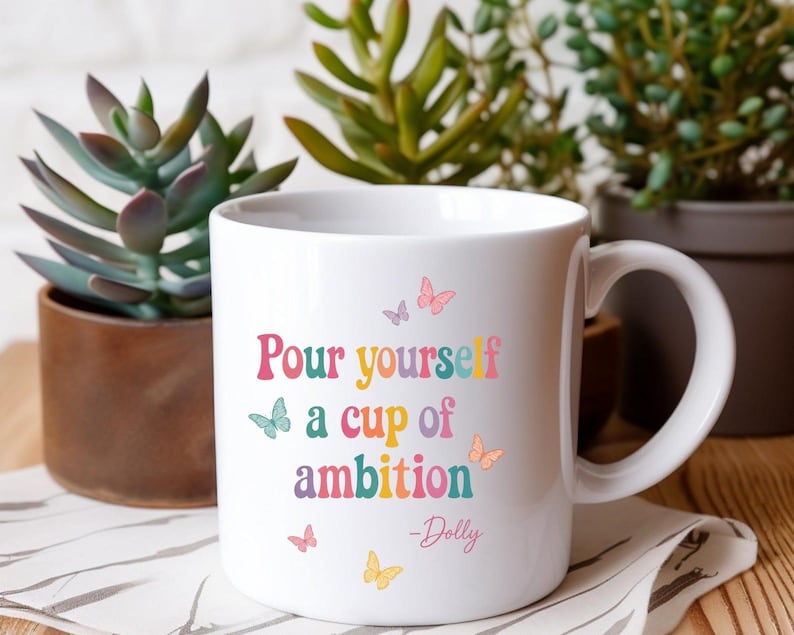 May include: A white ceramic mug featuring the colorful text "Pour yourself a cup of ambition" followed by "-Dolly" in a script font. The design is decorated with small, multi-colored butterflies in shades of pink, yellow, blue, and purple.
