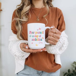 May include: A white ceramic mug featuring the colorful text Pour yourself a cup of ambition and the signature -Dolly, accented with small pastel-colored butterfly illustrations in pink, blue, orange, and yellow scattered around the lettering.