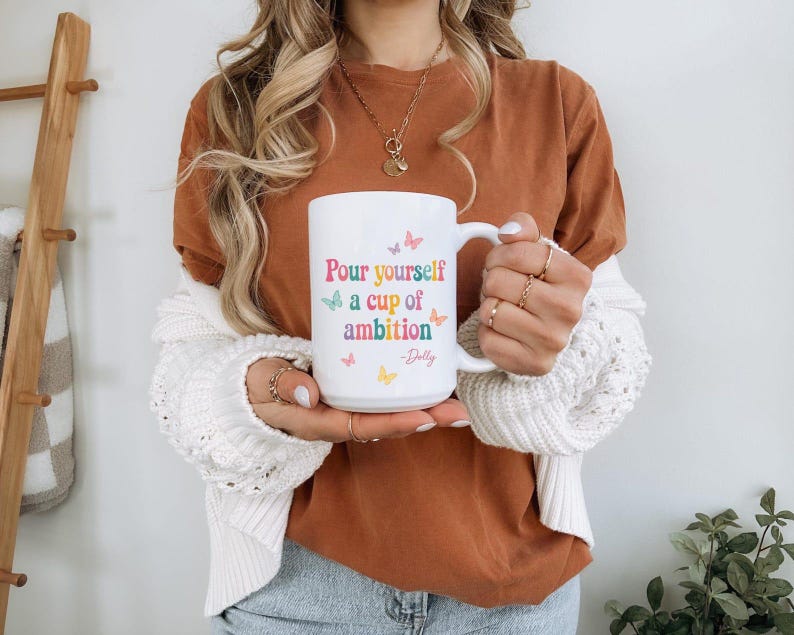 May include: A white ceramic mug featuring the colorful text Pour yourself a cup of ambition and the signature -Dolly, accented with small pastel-colored butterfly illustrations in pink, blue, orange, and yellow scattered around the lettering.