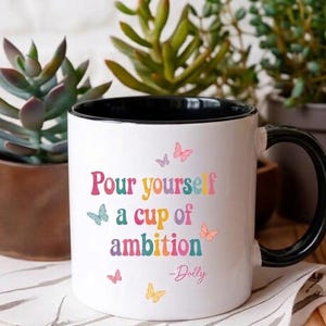 May include: A white ceramic mug with a black interior and handle. The front features the colorful text "Pour yourself a cup of ambition" followed by "-Dolly" in a script font. Small, multi-colored butterfly illustrations surround the text.