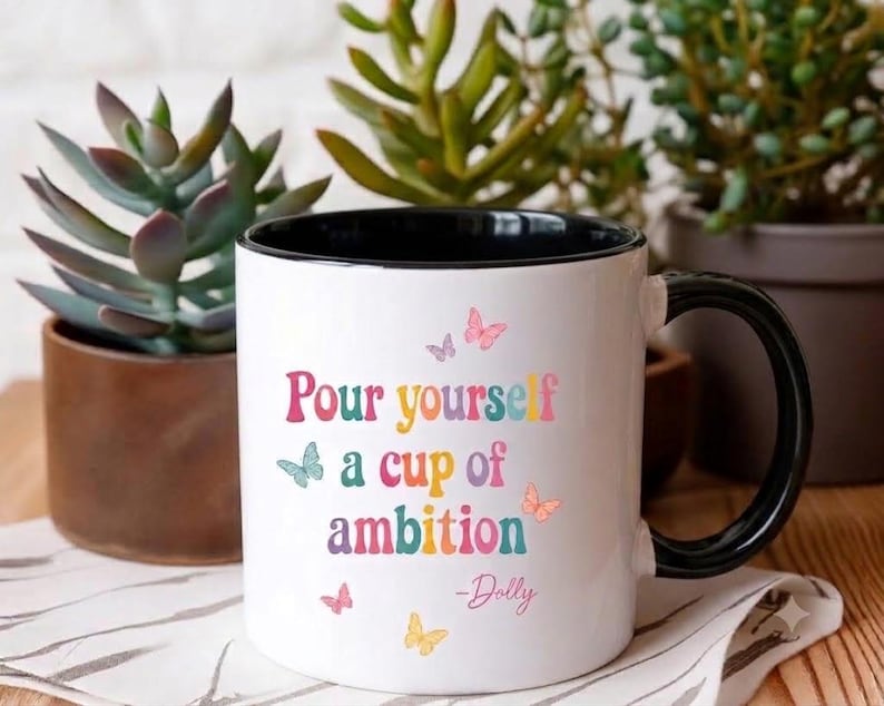 May include: A white ceramic mug with a black interior and handle. The front features the colorful text "Pour yourself a cup of ambition" followed by "-Dolly" in a script font. Small, multi-colored butterfly illustrations surround the text.