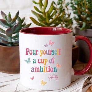 May include: A white ceramic mug with a deep red interior and handle. The front features the colorful text "Pour yourself a cup of ambition" followed by "-Dolly" in a script font, accented by small, multi-colored butterfly illustrations.