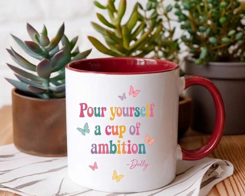 May include: A white ceramic mug with a deep red interior and handle. The front features the colorful text "Pour yourself a cup of ambition" followed by "-Dolly" in a script font, accented by small, multi-colored butterfly illustrations.