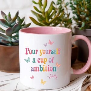 May include: A white ceramic mug with a light pink interior and handle. The front features the colorful text "Pour yourself a cup of ambition -Dolly" in a retro font, surrounded by small, multi-colored butterfly illustrations in pink, blue, and yellow.