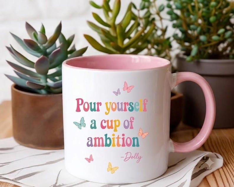 May include: A white ceramic mug with a light pink interior and handle. The front features the colorful text "Pour yourself a cup of ambition -Dolly" in a retro font, surrounded by small, multi-colored butterfly illustrations in pink, blue, and yellow.