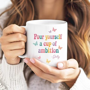 May include: A white ceramic mug featuring the colorful text Pour yourself a cup of ambition -Dolly. The design includes small butterflies in shades of pink, blue, and yellow scattered around the lettering on the front of the mug.