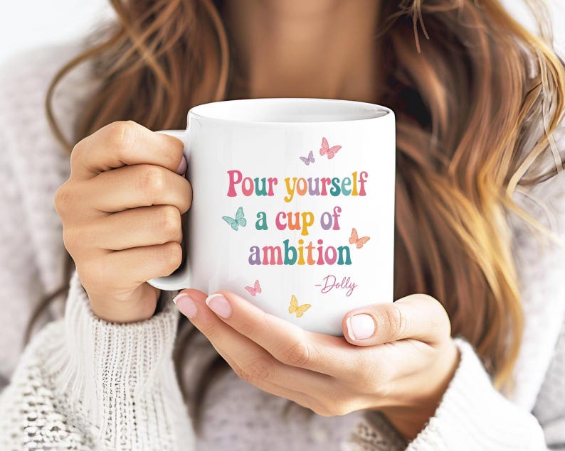 May include: A white ceramic mug featuring the colorful text Pour yourself a cup of ambition -Dolly. The design includes small butterflies in shades of pink, blue, and yellow scattered around the lettering on the front of the mug.