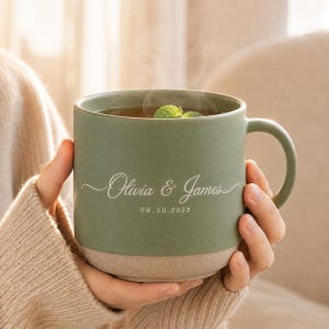 Peut inclure: Un mug en céramique vert sauge avec une finition mate et une base beige mouchetée. L'avant présente le texte blanc en cursive Olivia & James au-dessus de la date 08.30.2025. De la vapeur s'échappe du mug contenant un brin de menthe verte.