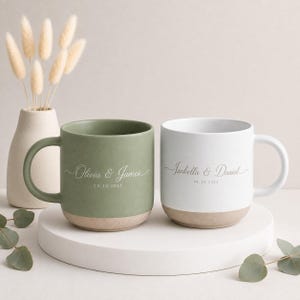May include: Two ceramic mugs with a matte finish, one sage green and one white, each featuring a tan base. The green mug is printed with the text Olivia & James 08.30.2025, and the white mug is printed with the text Isabella & Daniel 09.30.2025.