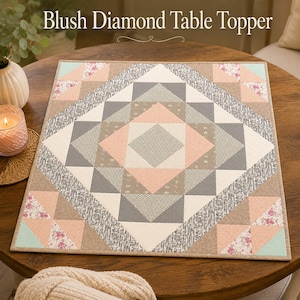 May include: A square fabric table topper titled Blush Diamond Table Topper featuring a geometric quilt pattern in shades of blush pink, charcoal grey, cream, and beige with floral accents. The design consists of triangles and squares.