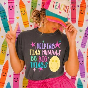 Op de afbeelding: Een antracietgrijs t-shirt met de kleurrijke tekst HELPING TINY HUMANS DO BIG THINGS en een geel smileygezicht. Het t-shirt wordt gecombineerd met een witte, roze en turquoise truckerpet met het woord TEACHER in grote, kleurrijke letters.