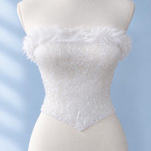 May include: A strapless white corset top featuring a textured, shimmering fabric throughout the bodice and a soft, fluffy white faux fur trim along the upper neckline. The garment has a pointed hemline at the center front.