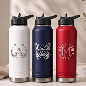 May include: Three stainless steel water bottles with black lids and straws. The white bottle features a laurel wreath and the name AMELIA. The navy blue bottle displays a butterfly graphic and the name LUANA. The red bottle shows a letter M.
