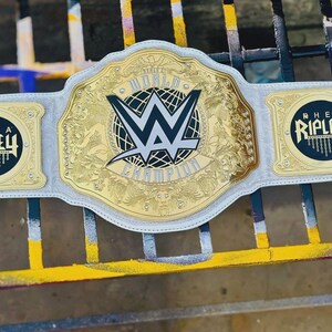 May include: A white leather championship wrestling belt featuring a large central gold plate with the WWE logo and the text World Champion. Two smaller gold side plates feature the black and gold text Rhea Ripley with a chain graphic design.