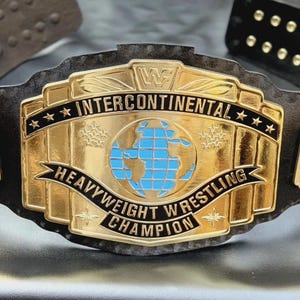 May include: A gold-colored championship wrestling belt featuring a central plate with a blue globe graphic and the text "INTERCONTINENTAL HEAVYWEIGHT WRESTLING CHAMPION" and the "WWF" logo. The belt has a black leather strap with gold metal studs.