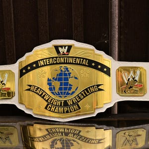 May include: A white championship wrestling belt featuring a large central gold plate with a blue globe graphic and the text INTERCONTINENTAL HEAVYWEIGHT WRESTLING CHAMPION. Four smaller gold side plates depict wrestling figures and the WWE logo.