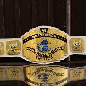 May include: A white leather wrestling championship belt featuring a large central gold plate with a blue globe graphic and the text INTERCONTINENTAL HEAVYWEIGHT WRESTLING CHAMPION. The belt includes four smaller gold side plates with wrestling figures.
