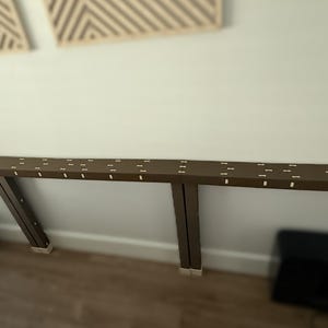 May include: A long, narrow, brown horizontal shelf or console table mounted to a wall. The top surface features a repeating pattern of small, light-colored rectangular inlays and two circular cutouts, one located at each far end of the structure.
