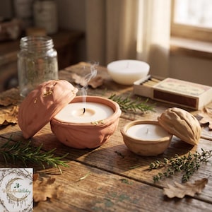 Op de afbeelding: Twee terracotta-kleurige kaarsenhouders in de vorm van walnootschalen, waarvan één met een brandende kaars met een dunne rookpluim en bladgoudaccenten, en de andere met een kleine witte kaars. De set bevat een doosje lucifers en wat groen.
