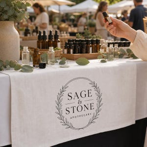 Puede incluir: Un camino de mesa de lino blanco con un logotipo circular negro que presenta un diseño de corona de hojas y el texto SAGE & STONE APOTHECARY. Se exhibe sobre una mesa con varios frascos cuentagotas de vidrio ámbar y un jarrón de cerámica.