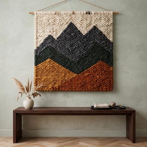 Könnte beinhalten: Ein quadratischer Wandbehang aus dickem, strukturiertem Garn mit einem Berglandschaftsmotiv. Der Wandteppich zeigt horizontale Farbblöcke in Creme, Dunkelanthrazit und gebranntem Orange und ist an einer Holzstange mit einer Kordel aufgehängt.