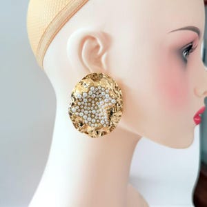 May include: A large, circular gold-tone earring with an irregular, hammered metal surface. The center of the earring features a dense cluster of small, round white faux pearls arranged in a textured, organic pattern across the metallic base.
