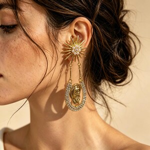 May include: A gold-toned statement earring featuring a sunburst top adorned with small white pearls. Below the sunburst, a U-shaped pendant encrusted with clear crystals surrounds a central gold-toned sculptural element shaped like a human ear.