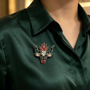 May include: A gold-toned metal brooch shaped like a bull head, featuring red, teal, and black enamel detailing. The design includes a central heart-shaped clear crystal on the forehead and two smaller oval clear crystals set into the bull's eyes.