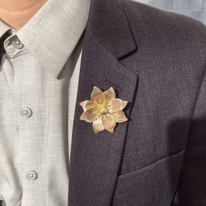 May include: A gold-toned metal brooch shaped like a multi-petaled flower with fine linear texturing on each petal. The brooch is pinned to the lapel of a dark charcoal gray suit jacket worn over a light gray button-down collared shirt.