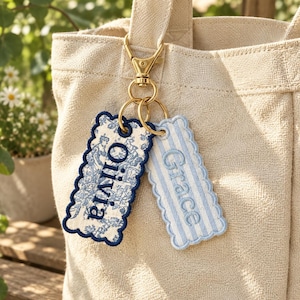 May include: Two rectangular fabric bag tags with scalloped edges attached to a gold-tone metal lobster clasp. One tag features a blue and white floral toile pattern with the name Olivia, and the other has blue and white stripes with the name Grace.