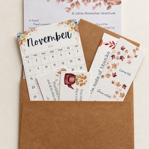 May include: A brown paper envelope containing a November 2026 calendar, a card titled A Little November Gratitude, a card with a teapot illustration, and a bookmark with falling leaves and the text Leaves fall and Stories unfold.