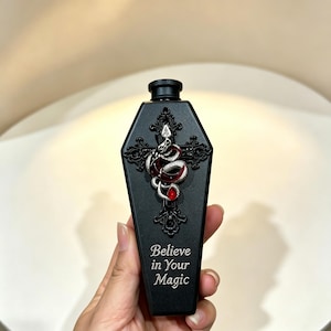 Pode incluir: Uma garrafa preta fosca em forma de caixão com um emblema de cruz preta em relevo adornado com uma cobra prateada e uma gema vermelha. O texto "Believe in Your Magic" está impresso em letras brancas na parte inferior frontal da garrafa.