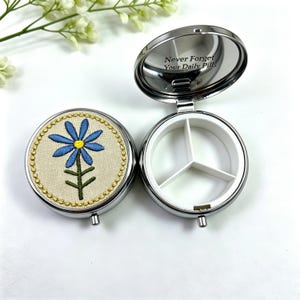 Puede incluir: Un pastillero redondo de plata con tapa abatible que presenta una flor azul bordada sobre una incrustación de tela beige. El interior es de plástico blanco con tres compartimentos. La tapa interior tiene grabado el texto Never Forget Your Daily Pills.