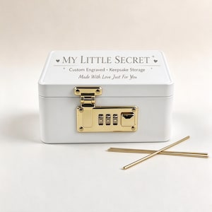 Pode incluir: Uma caixa de armazenamento retangular branca com um cadeado de combinação dourado na frente. A tampa está gravada com o texto MY LITTLE SECRET, Custom Engraved • Keepsake Storage e Made With Love Just For You. Duas hastes metálicas douradas.