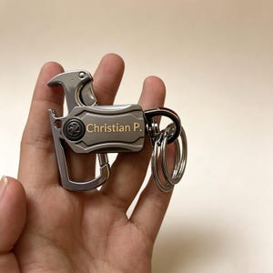 Puede incluir: Un llavero multiherramienta de color gris plomo con abrebotellas, mosquetón y dos anillas circulares. La placa rectangular central está grabada con el nombre Christian P. en una fuente de tono dorado sobre un acabado metálico.